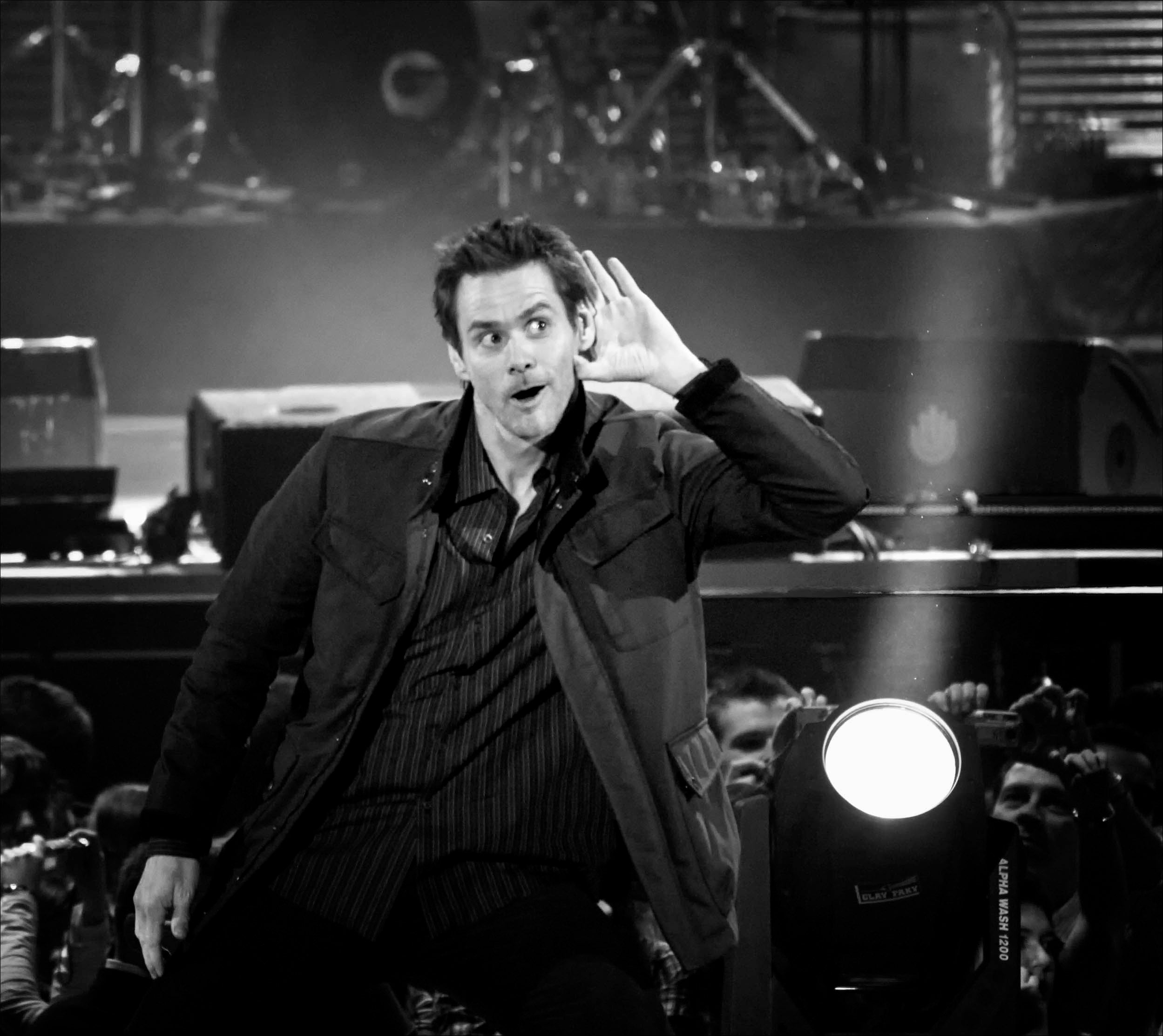 A man on stage holds his hand to his ear, appearing to listen to the audience. The photo is black and white with stage lights and equipment in the background.