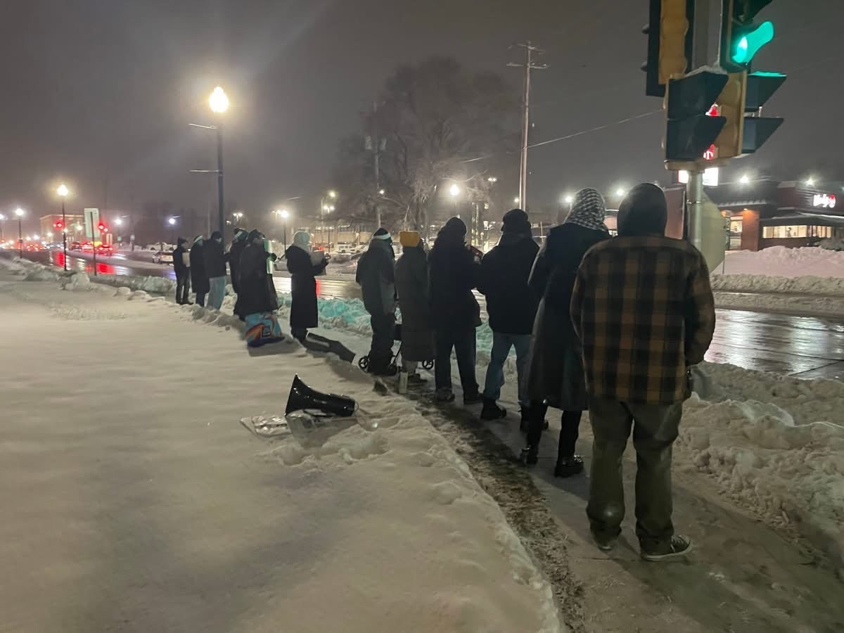 A group of people stand in a line on a snowy sidewalk at night near a traffic light, with snowbanks and wet roads visible.