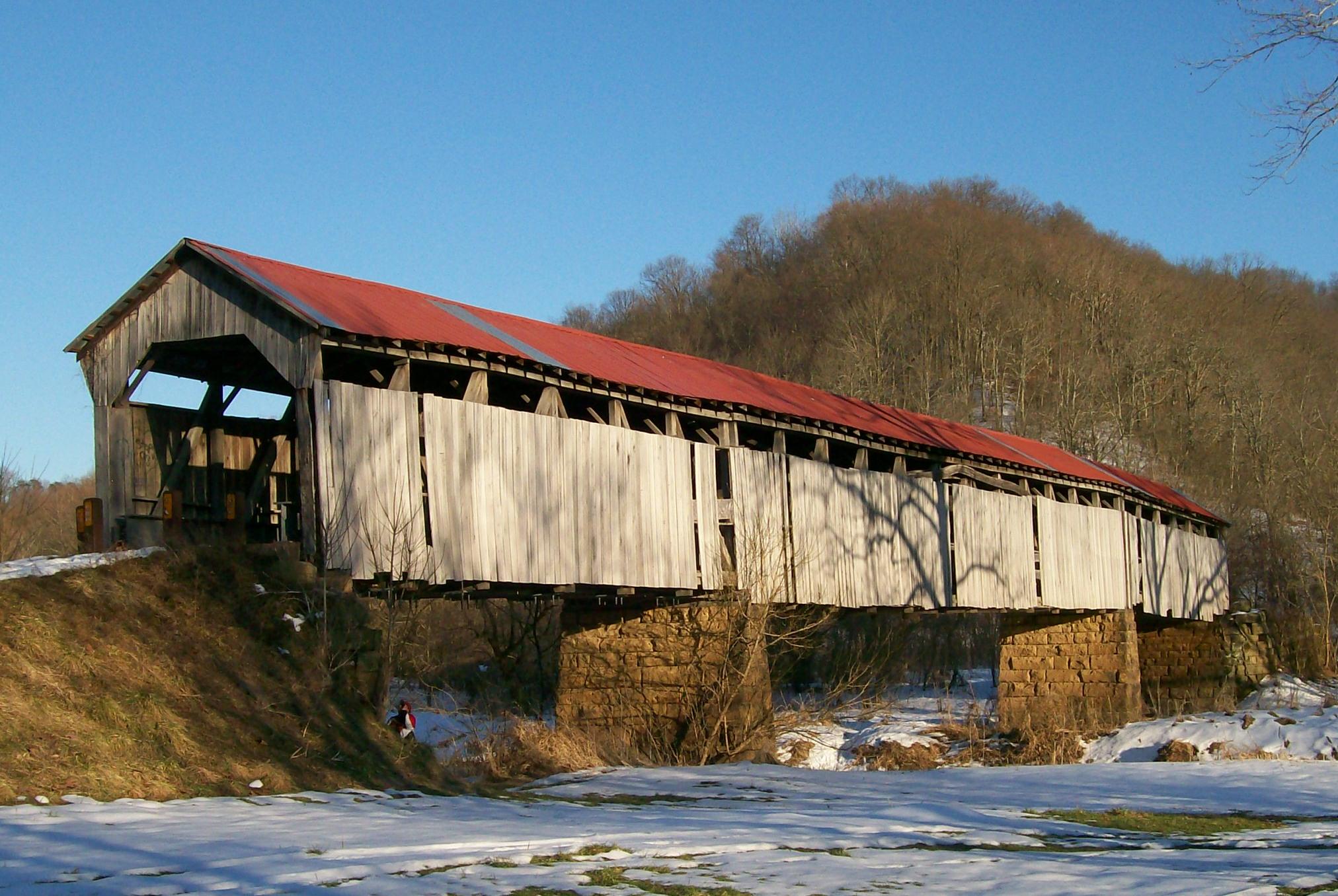 A long, wooden covered bridge with a red roof spans a partially snow-covered creek, supported by stone piers, with bare trees in the background.