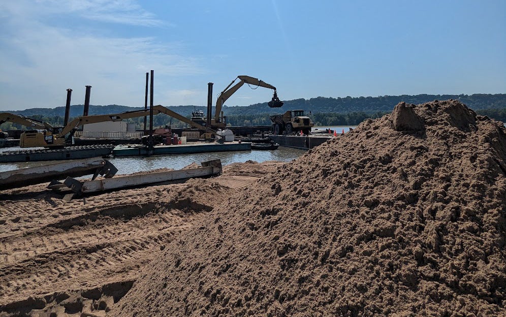 A large pile of sand sits near a riverbank, with construction equipment and barges visible on the water in the background under a clear sky.