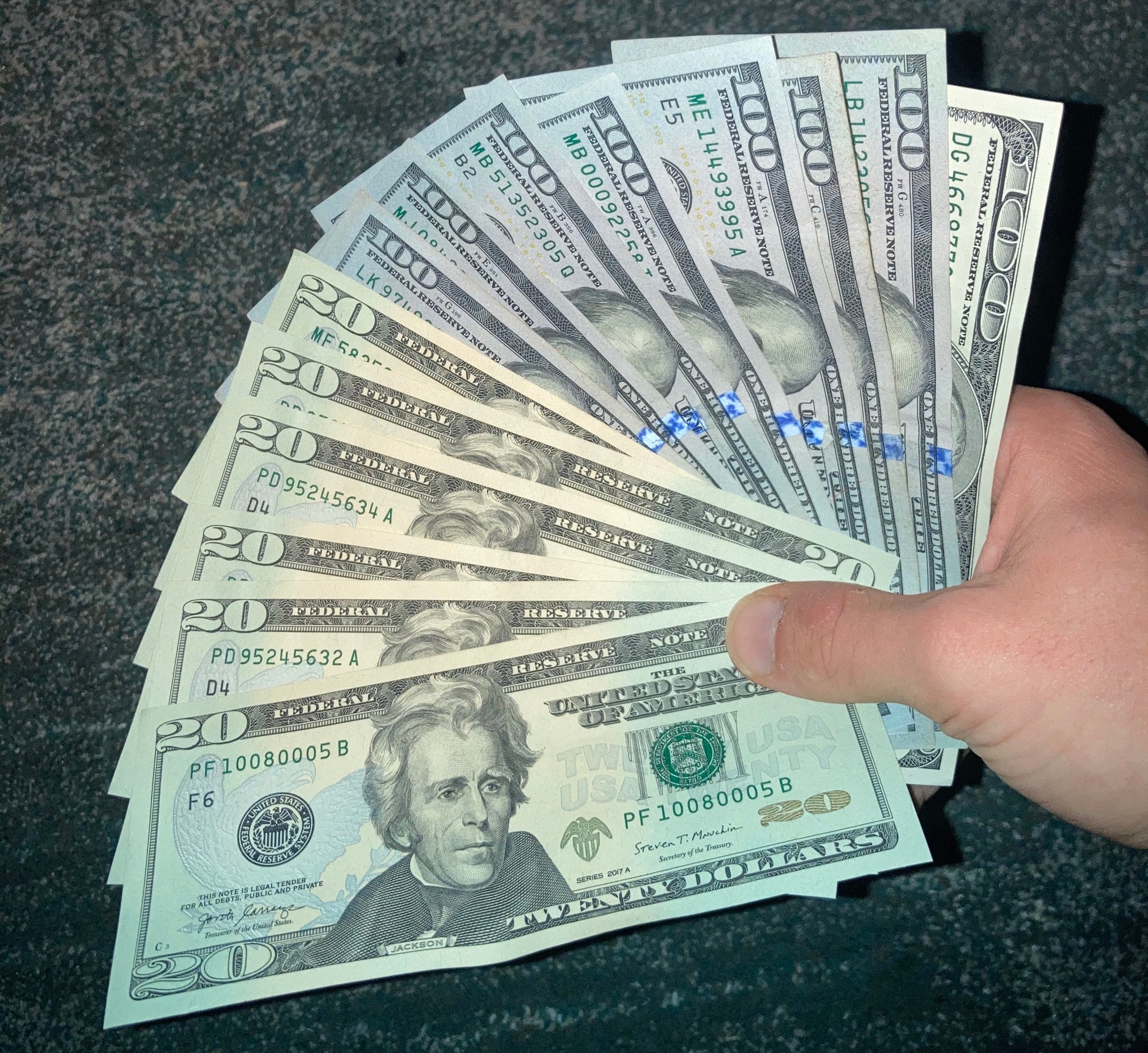 A hand holding a fanned-out stack of U.S. $100 and $20 bills against a dark background.
