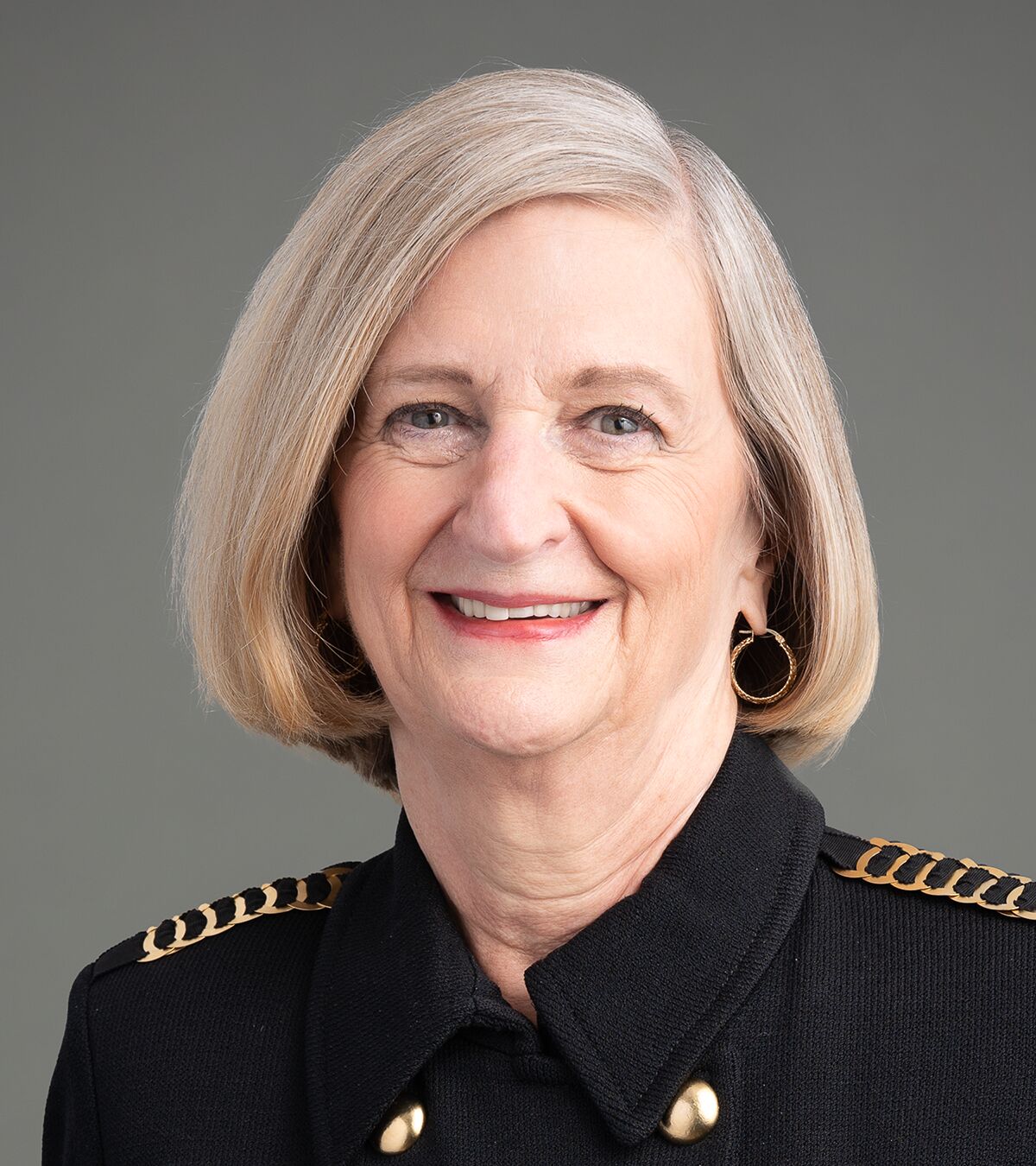 An older woman with short, gray hair smiles at the camera. She is wearing a black jacket with gold accents and hoop earrings, against a plain gray background.