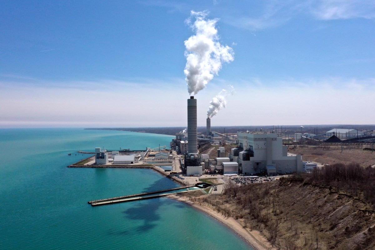 A large industrial power plant emits white smoke from tall stacks near a body of turquoise water, with clear skies overhead and some barren land in the foreground.
