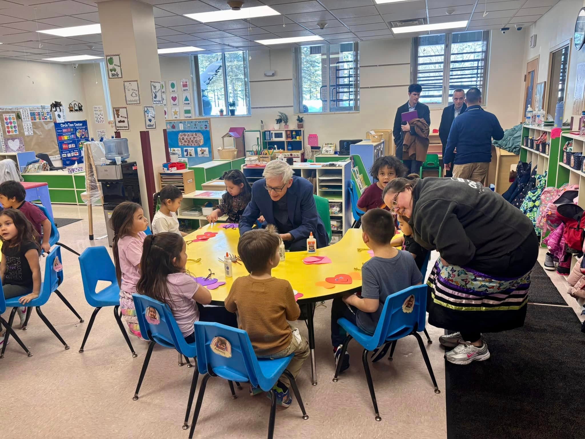 Adults observe and assist a group of young children seated around a table engaged in crafts in a brightly lit classroom.