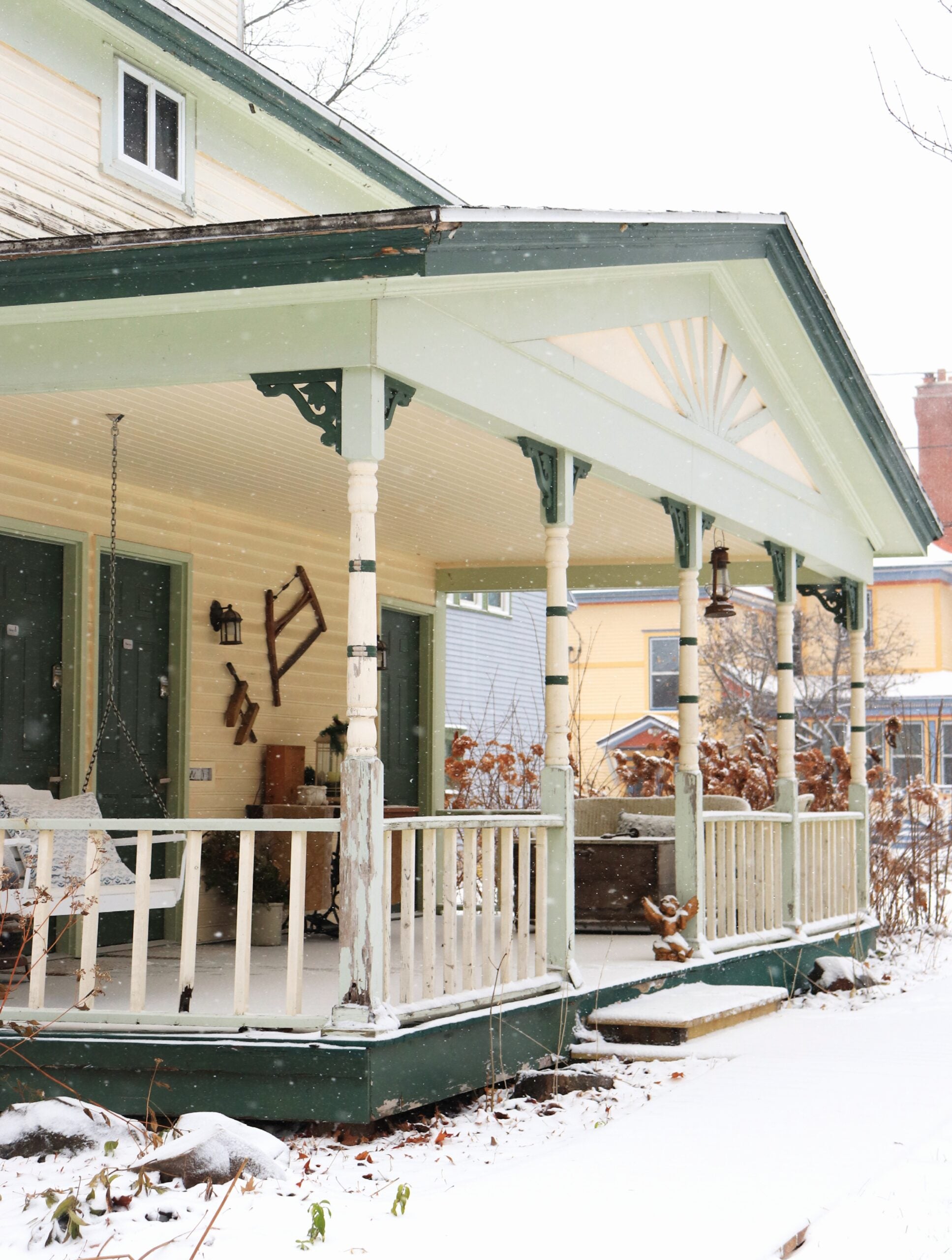A green and white house porch with a bench swing and a rocking chair, lightly dusted with snow in a winter setting.