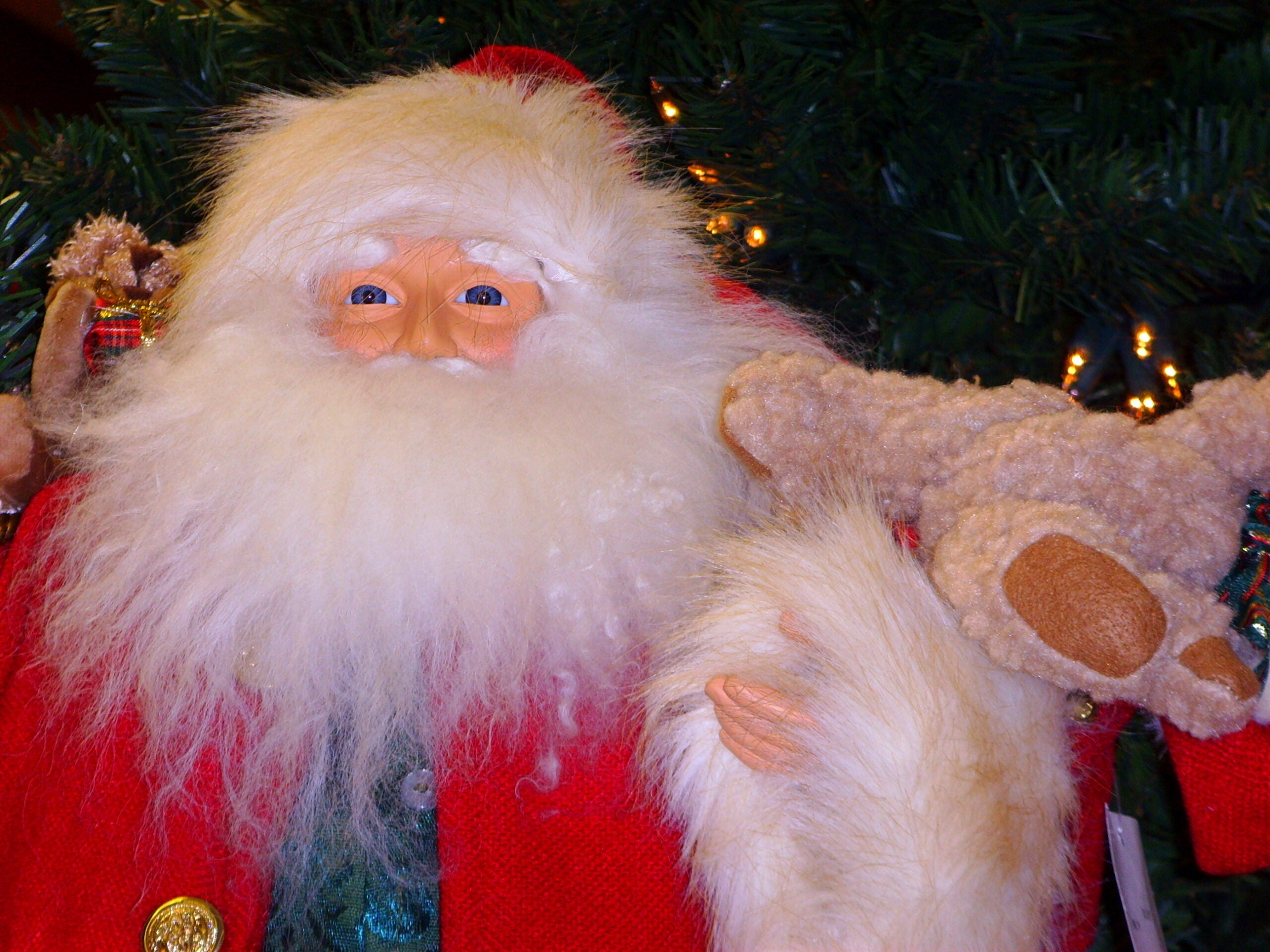 Santa Claus figurine with a white beard holds a tan stuffed teddy bear wearing a red coat, in front of a decorated Christmas tree.