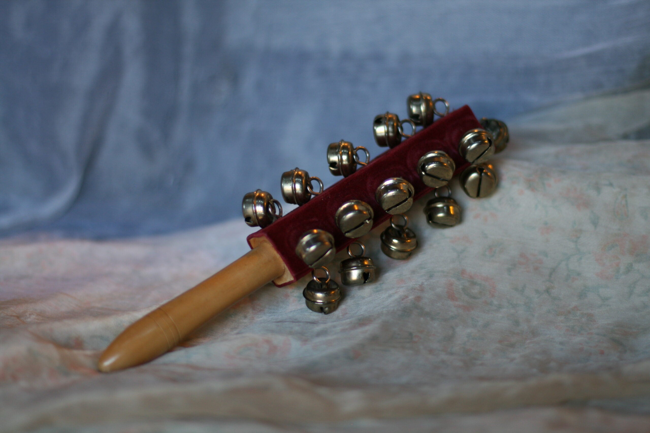 A wooden musical instrument with a handle and a velvet-covered bar featuring several metal jingle bells, placed on a patterned fabric surface.