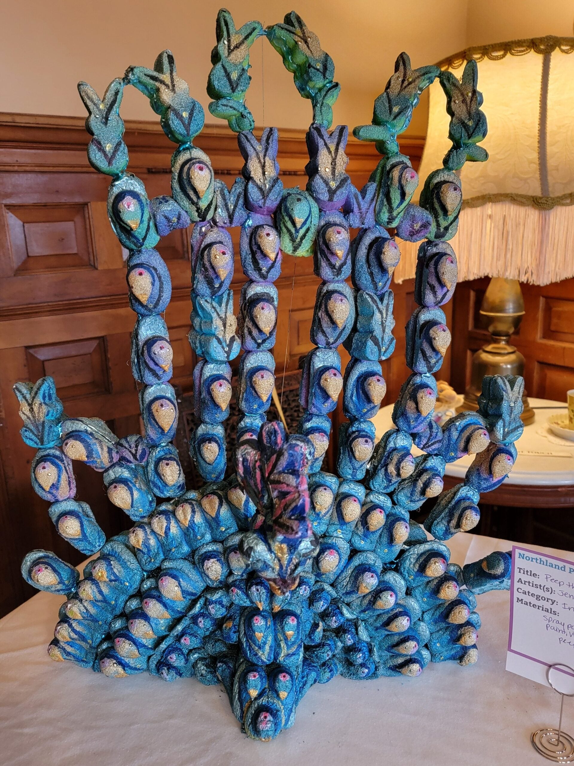 A colorful peacock sculpture made from painted pinecones is displayed on a table with a lamp and teacup in the background. A small placard with information is placed next to the sculpture.