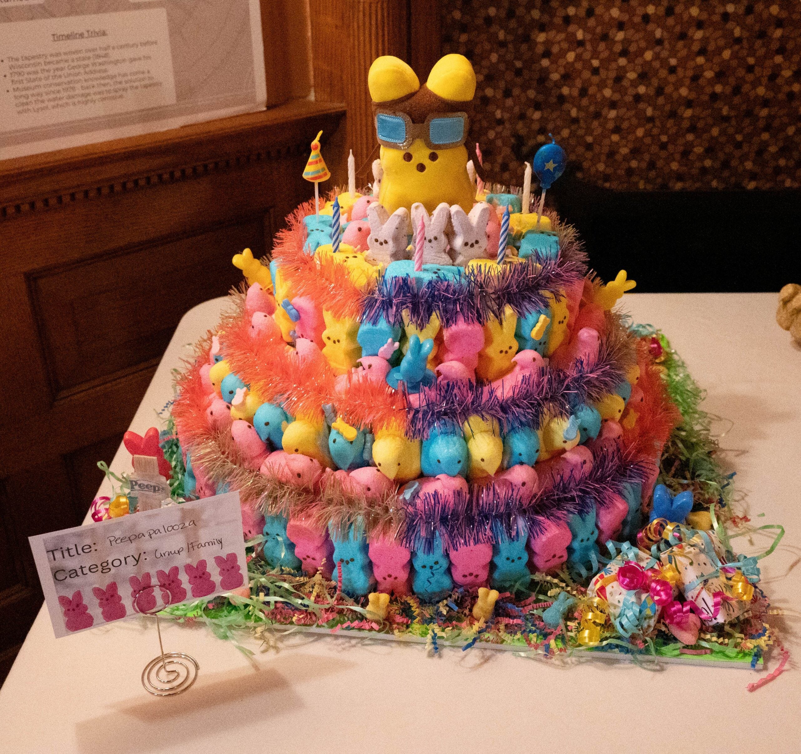 Three-tiered cake decorated with multicolored marshmallow Peeps, tinsel, and candy; a large yellow Peep with sunglasses and accessories sits on the top tier.