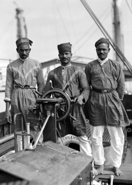 Three men wearing traditional attire stand on a boat, with one man at the wheel. The background shows rigging and parts of the ship.