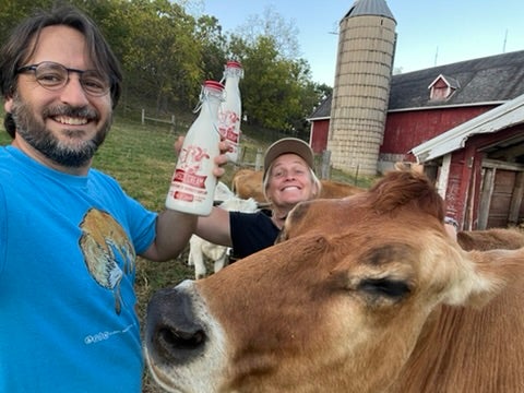 Two people stand next to a brown cow on a farm, holding glass bottles of milk. A red barn and a silo are visible in the background.