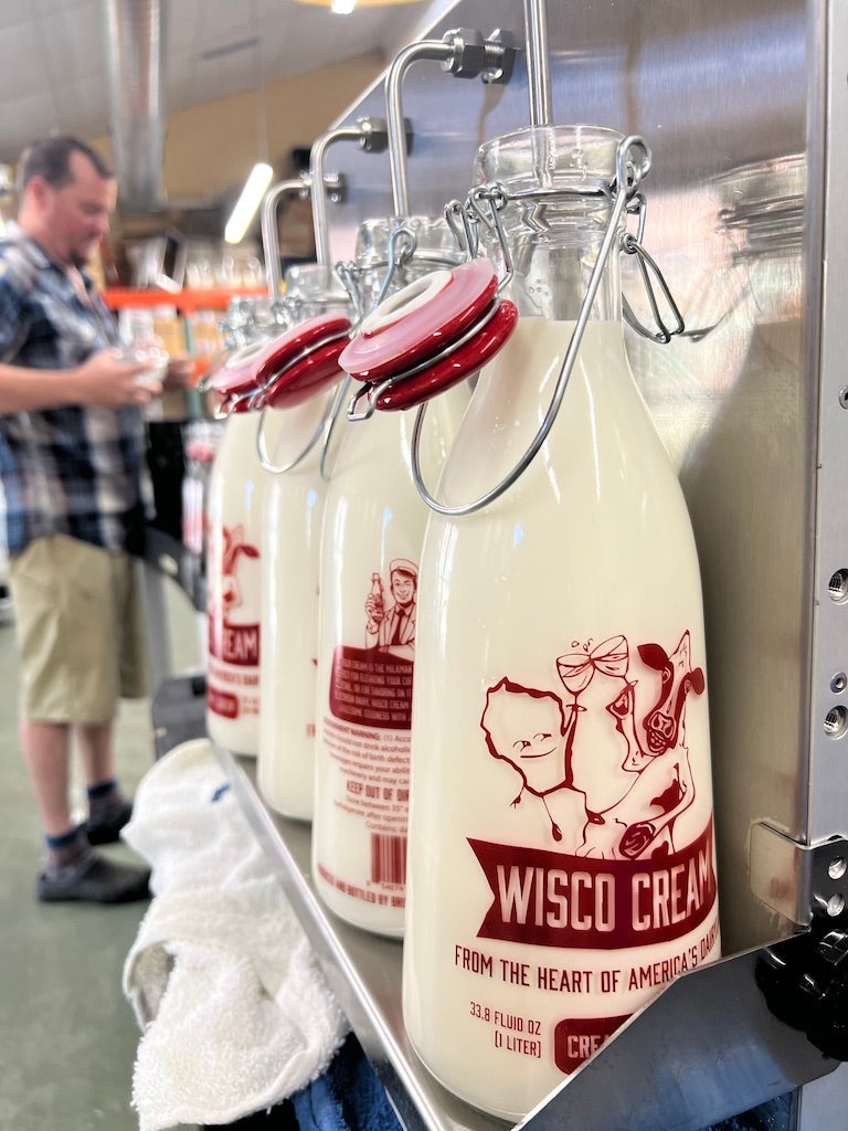 Several glass bottles of Wisco Cream milk with red labels are lined up on a counter; a man in a plaid shirt stands blurred in the background.