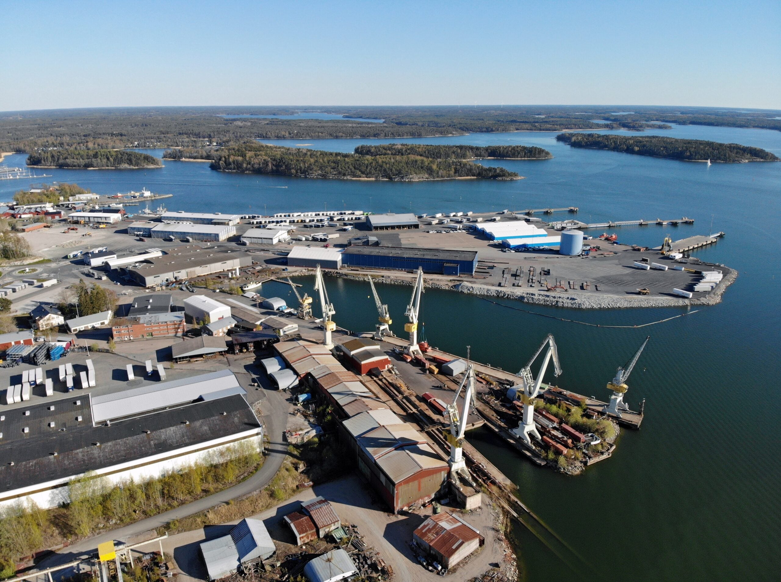 Aerial view of an industrial port with several large cranes, warehouses, docks, and cargo containers by a body of water, with forested islands in the background.
