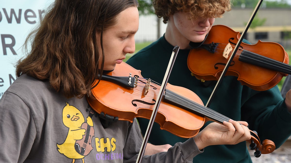 Two people play violins outdoors. The person in the foreground wears a gray shirt with a cartoon duck; the other in the background wears a green shirt.