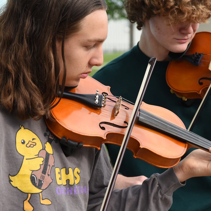 Two young people play violins outdoors. The person in front wears a gray shirt with a cartoon duck holding a violin and the text EHS ORCHESTRA.