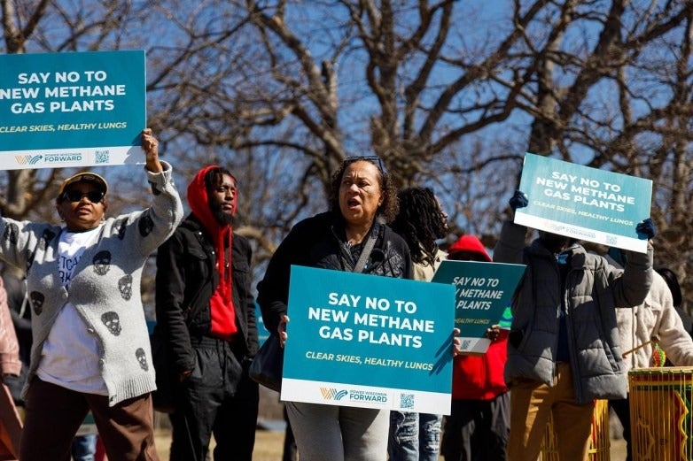 A group of people hold signs that read Say No to New Methane Gas Plants during an outdoor protest with leafless trees in the background.