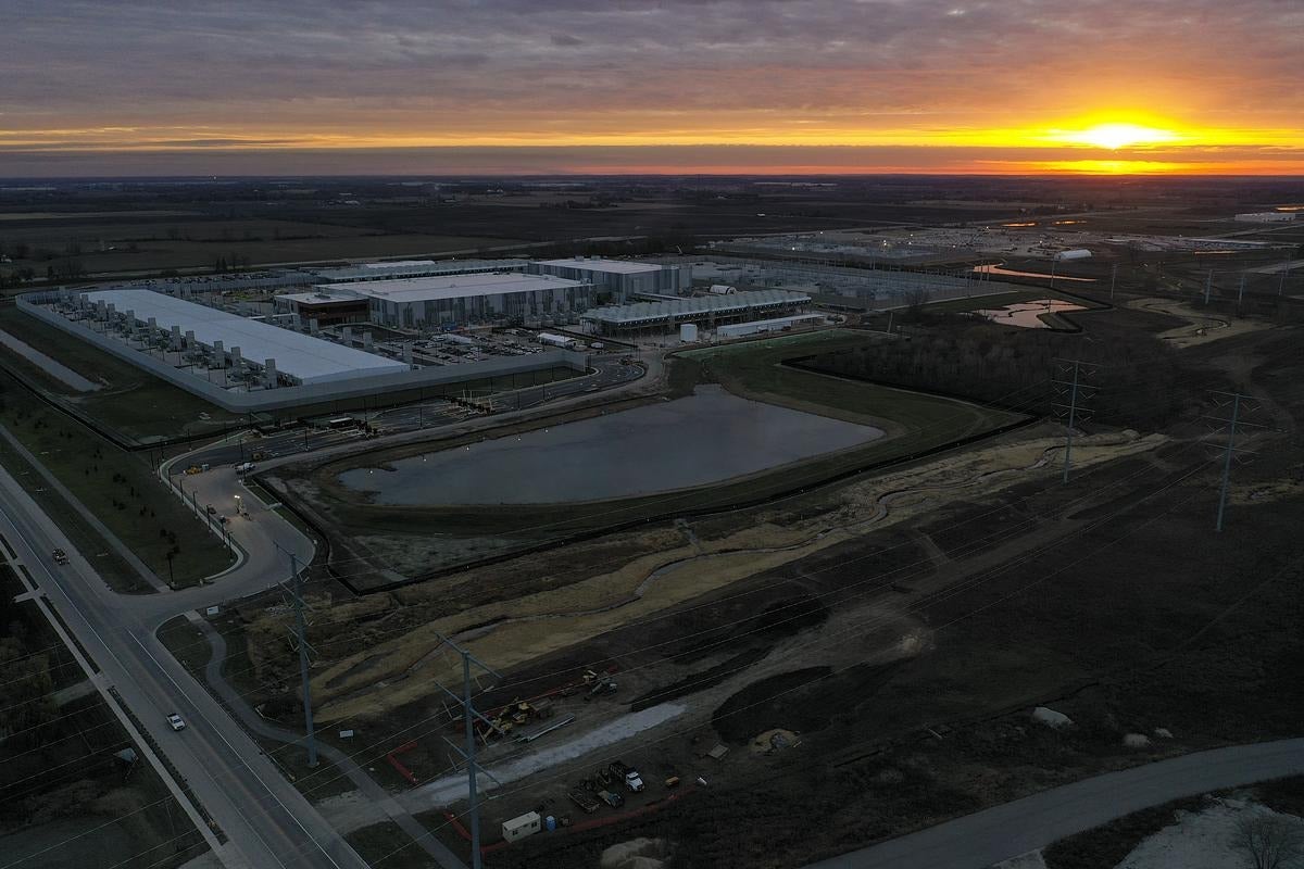 A large industrial facility with adjacent ponds and parking lots is shown at sunset, surrounded by roads and open fields under a partly cloudy sky.
