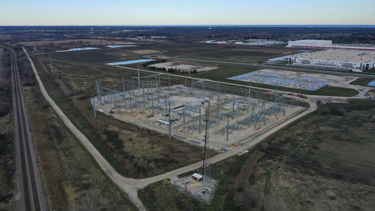 Aerial view of an electrical substation under construction near a warehouse and open fields, with a railway track running alongside the site.