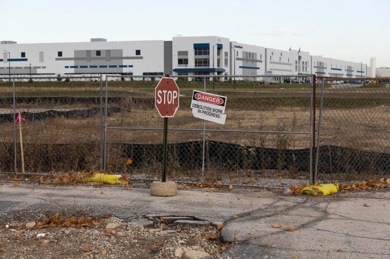 A stop sign and a Danger: Demolition Work in Progress sign are posted on a fence in front of a construction site with a large industrial building in the background.