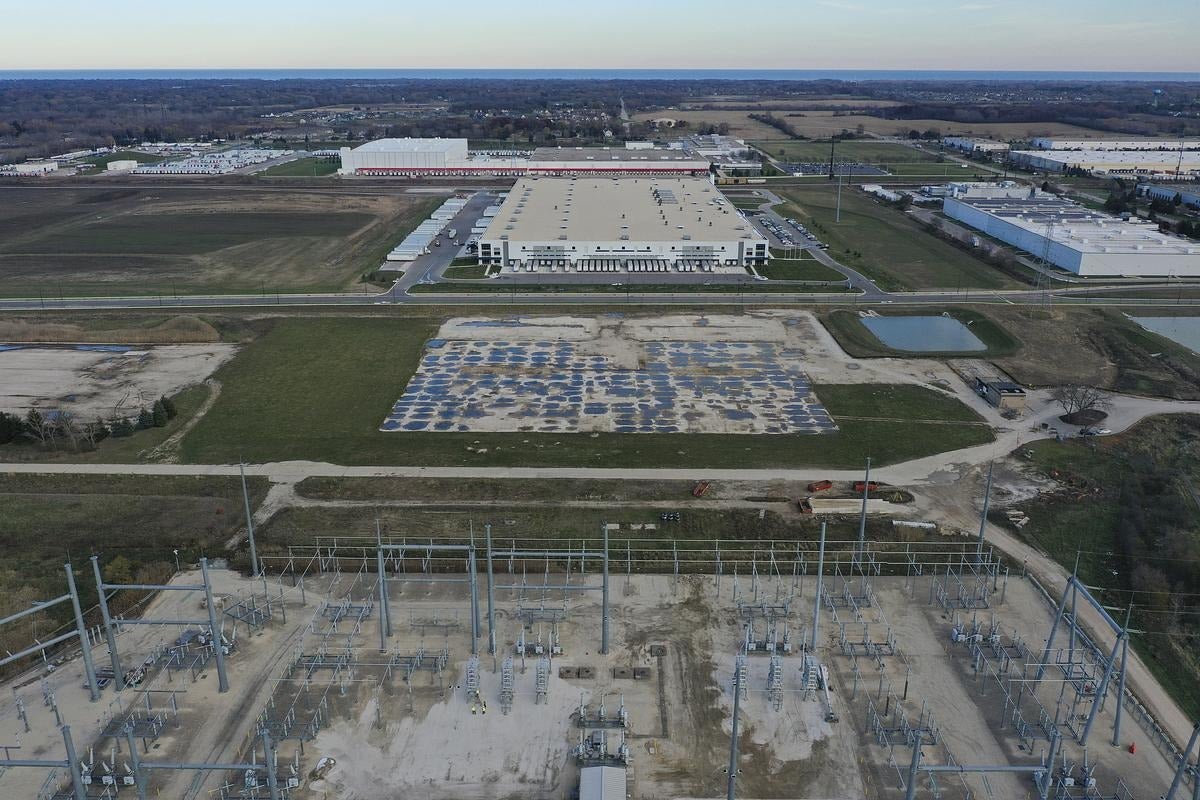 Aerial view of an industrial facility with a large warehouse, empty parking lot, open fields, and electrical substation in the foreground.