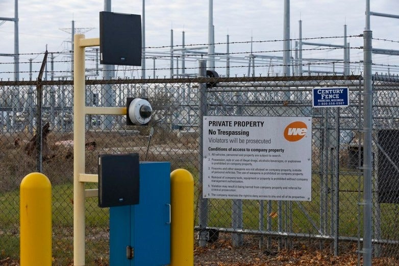 Chain-link fence with warning signs, surveillance camera, and security gate outside an industrial facility. Power lines and equipment are visible in the background.