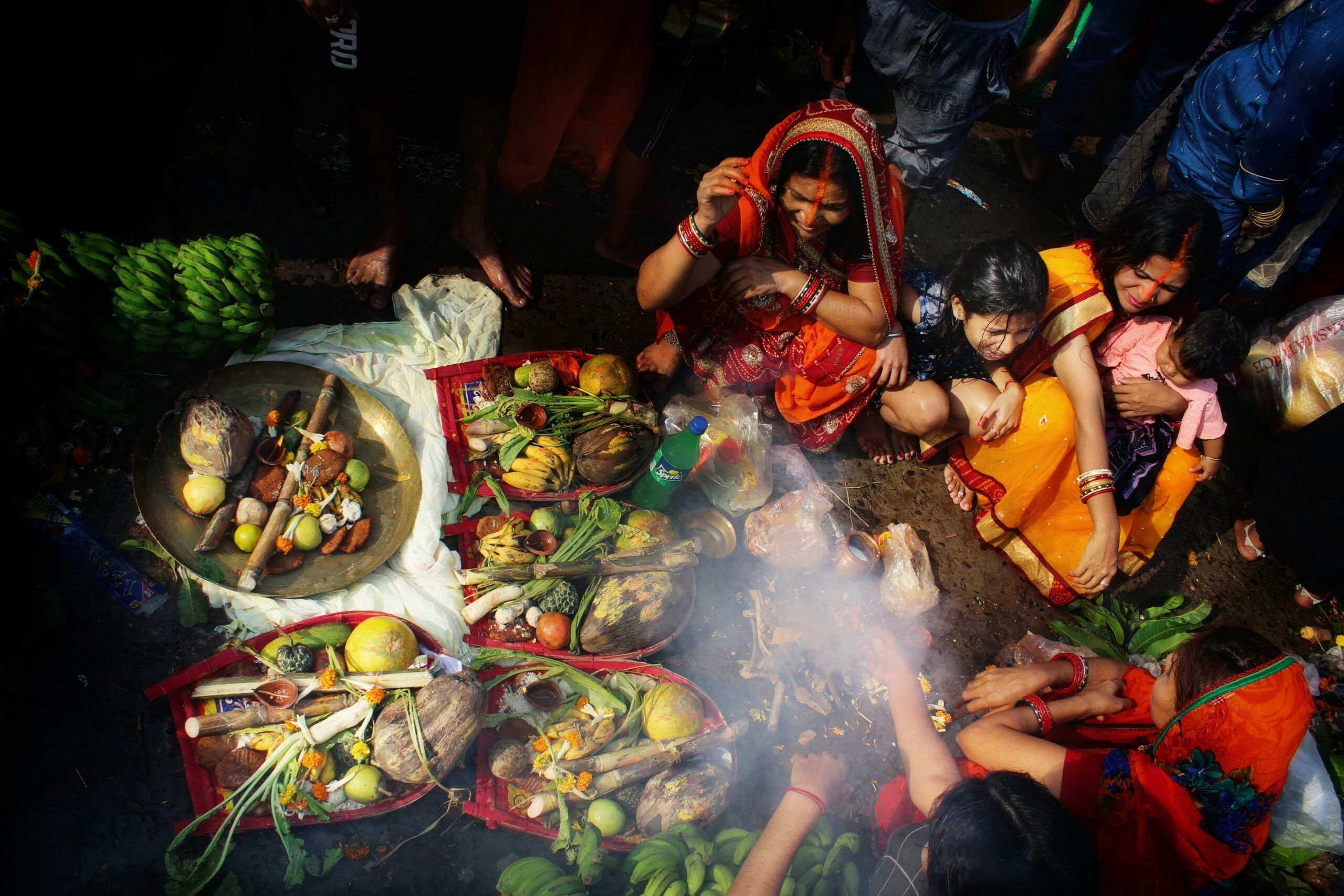 A group of women in colorful clothing perform a ritual with offerings of fruits and food items arranged in baskets, surrounded by smoke.