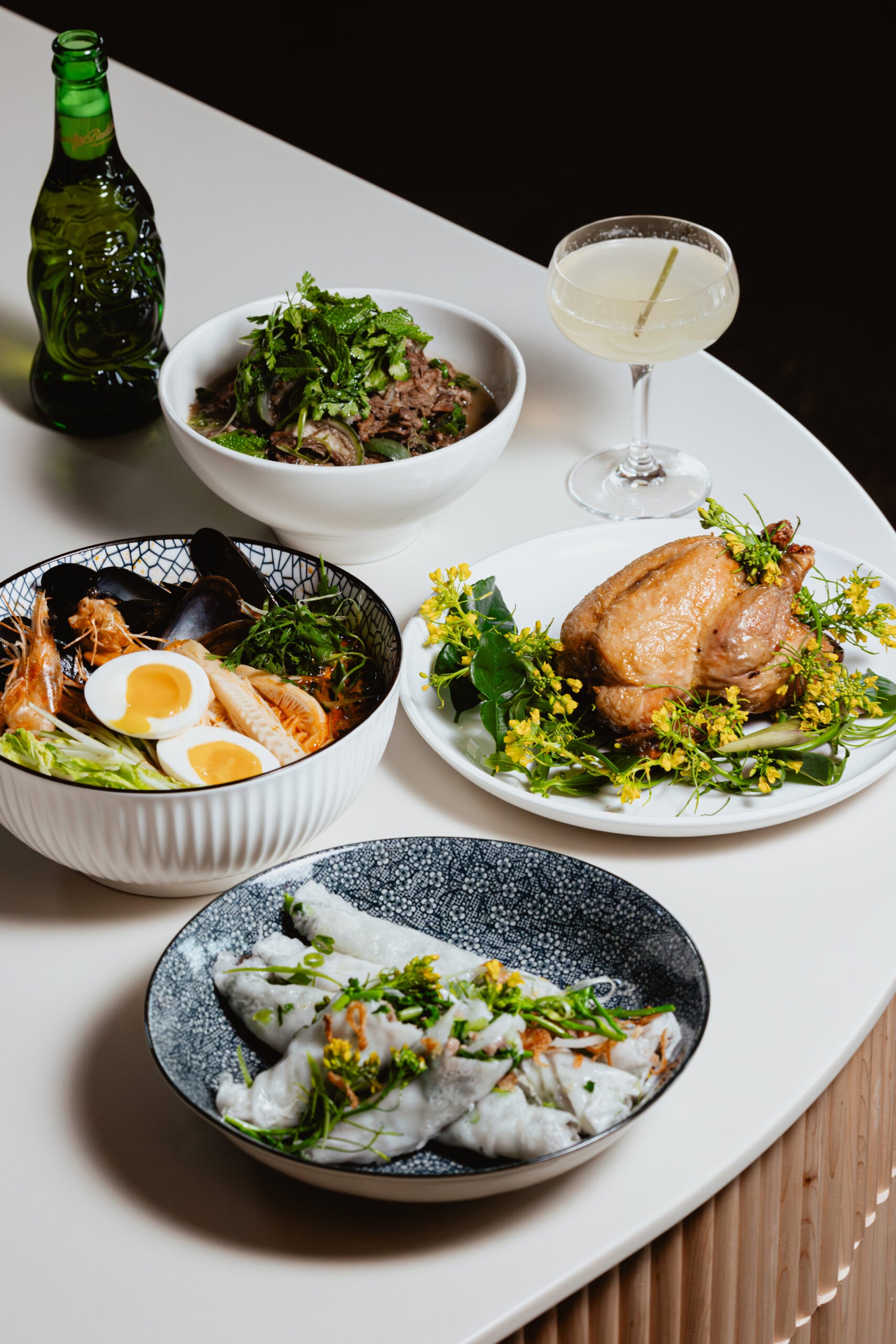 A table set with a green bottle, a cocktail, a bowl of noodles with herbs, a whole roasted chicken with greens, a bowl of ramen with eggs and seafood, and a plate of rice rolls with herbs.