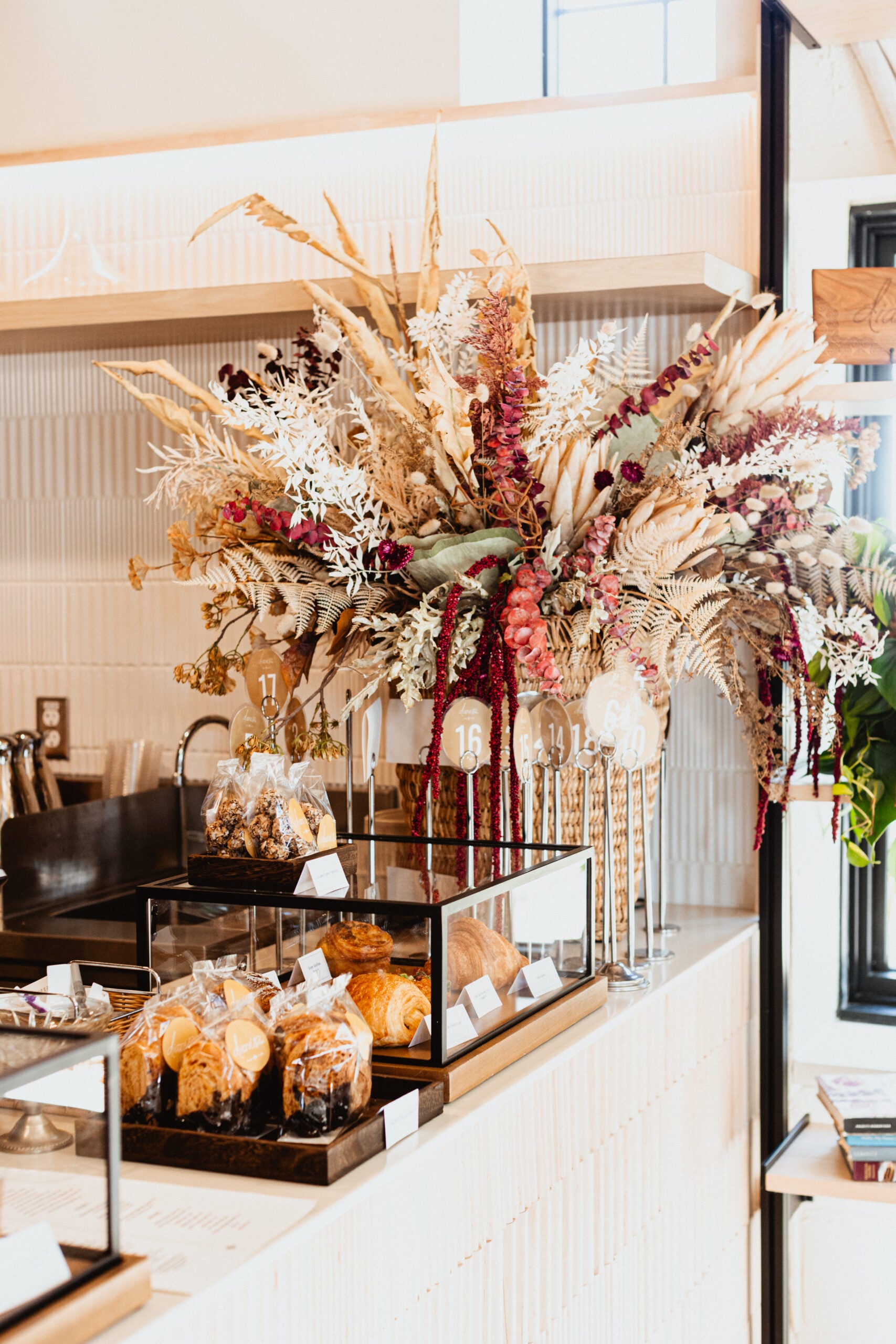 A bakery counter displays assorted pastries in glass cases with a large arrangement of dried flowers in vases behind the counter.