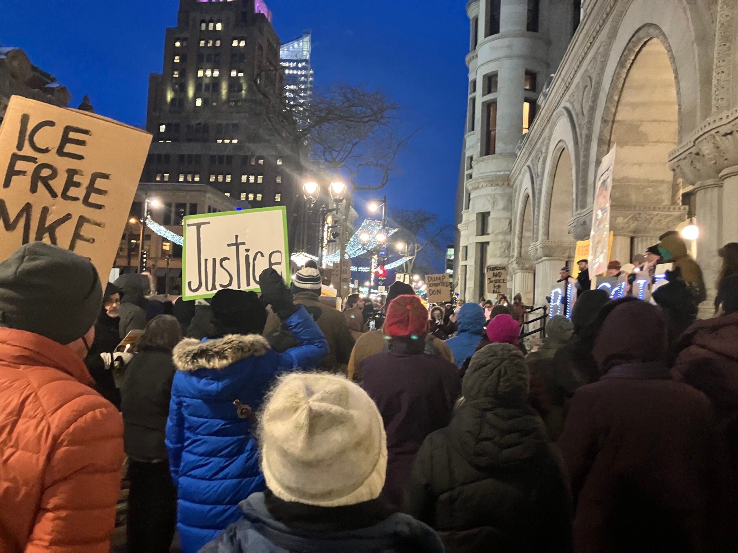 A crowd gathers outside a stone building at dusk, holding signs including one that says Justice and another that says ICE FREE MKE.