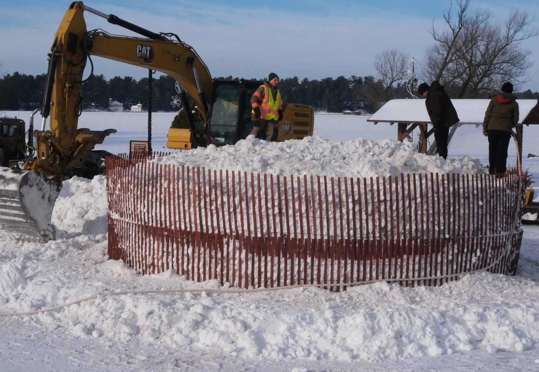 Workers use excavators to move snow into a fenced circular area outdoors in a snowy landscape, with trees and shelters in the background.