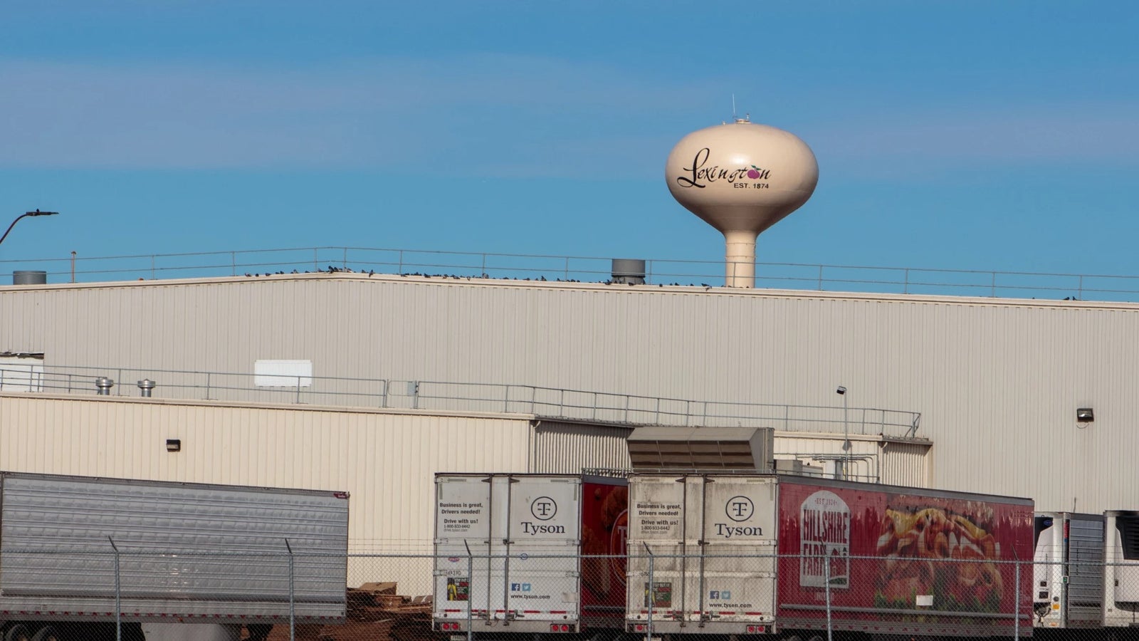 A beige industrial building with Tyson trucks parked outside and a water tower labeled Lexington in the background under a blue sky.