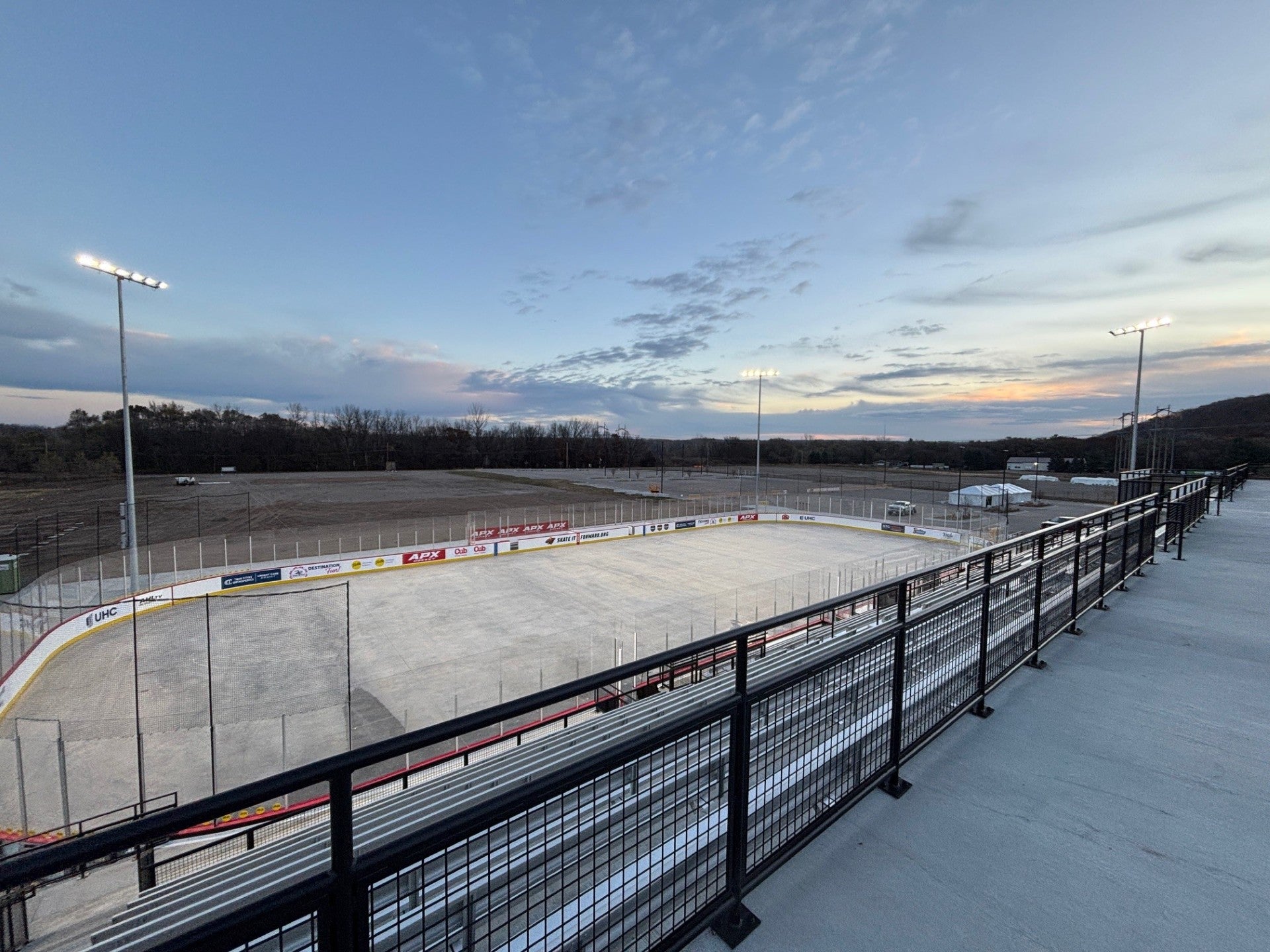 An empty outdoor hockey rink with surrounding bleachers and lit stadium lights at dusk, with a cloudy sky and trees in the background.