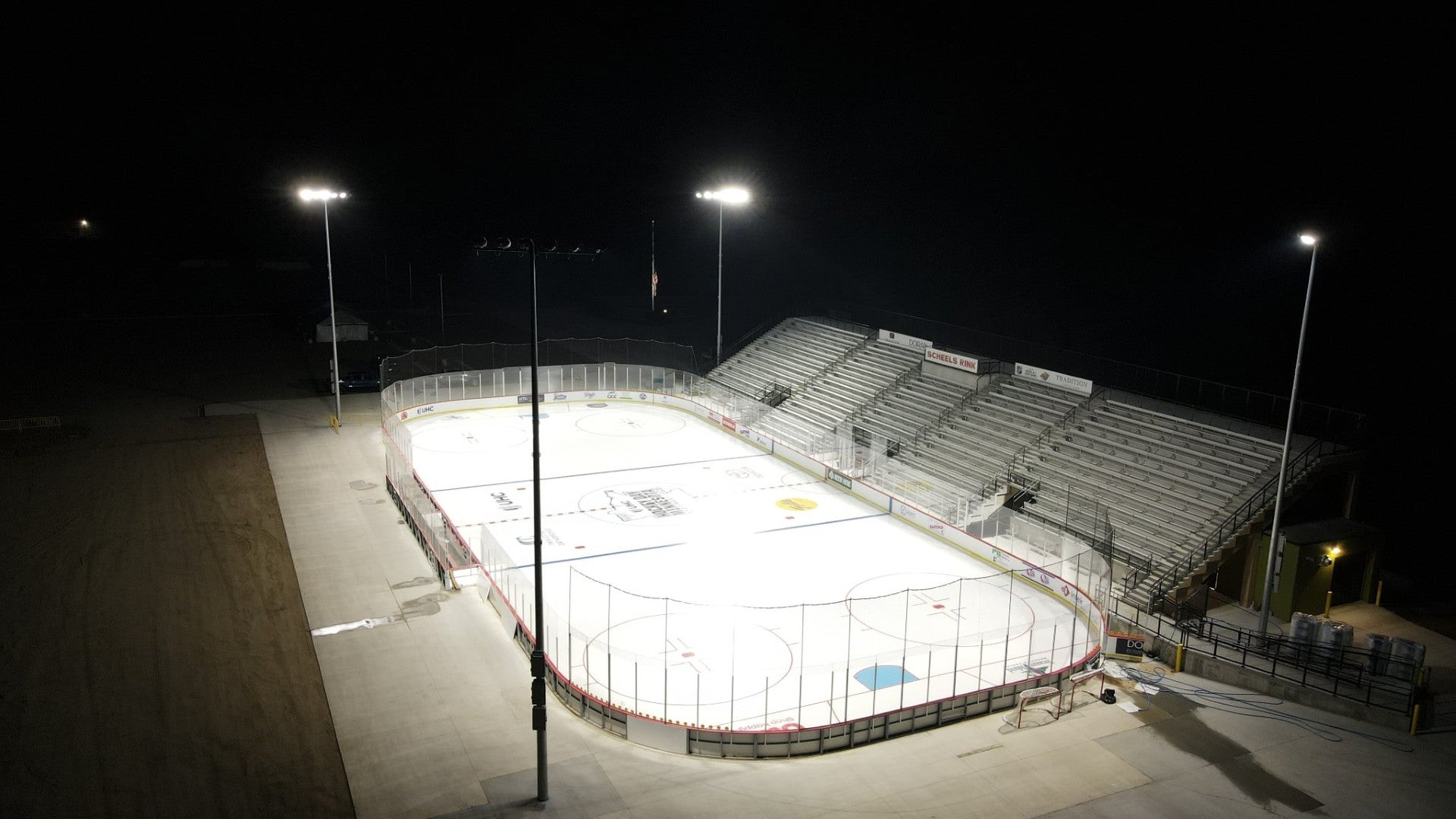 An outdoor hockey rink illuminated by floodlights at night, with empty bleachers on one side and no people present.