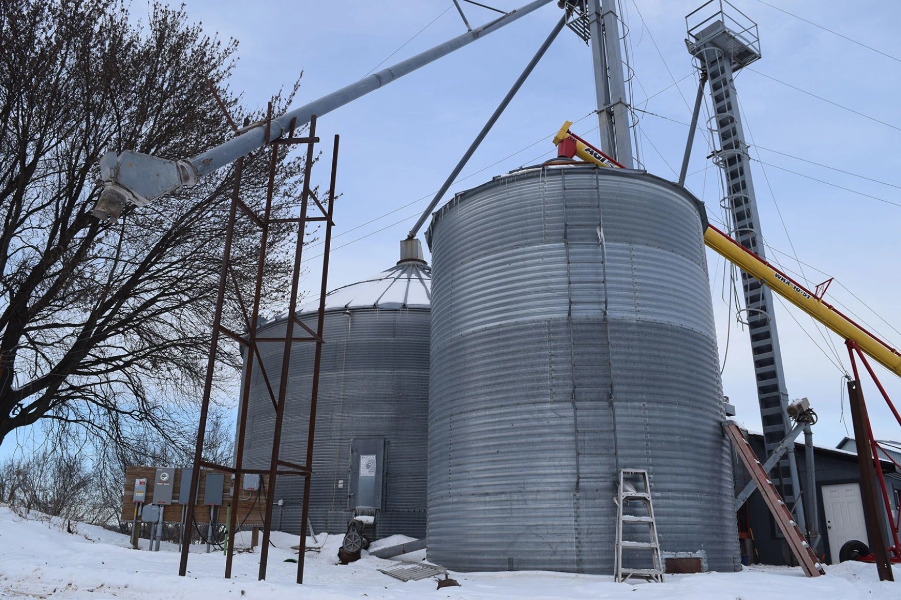 Two large metal grain silos and conveyor systems stand on snowy ground, with a ladder and some trees visible beside the structures.