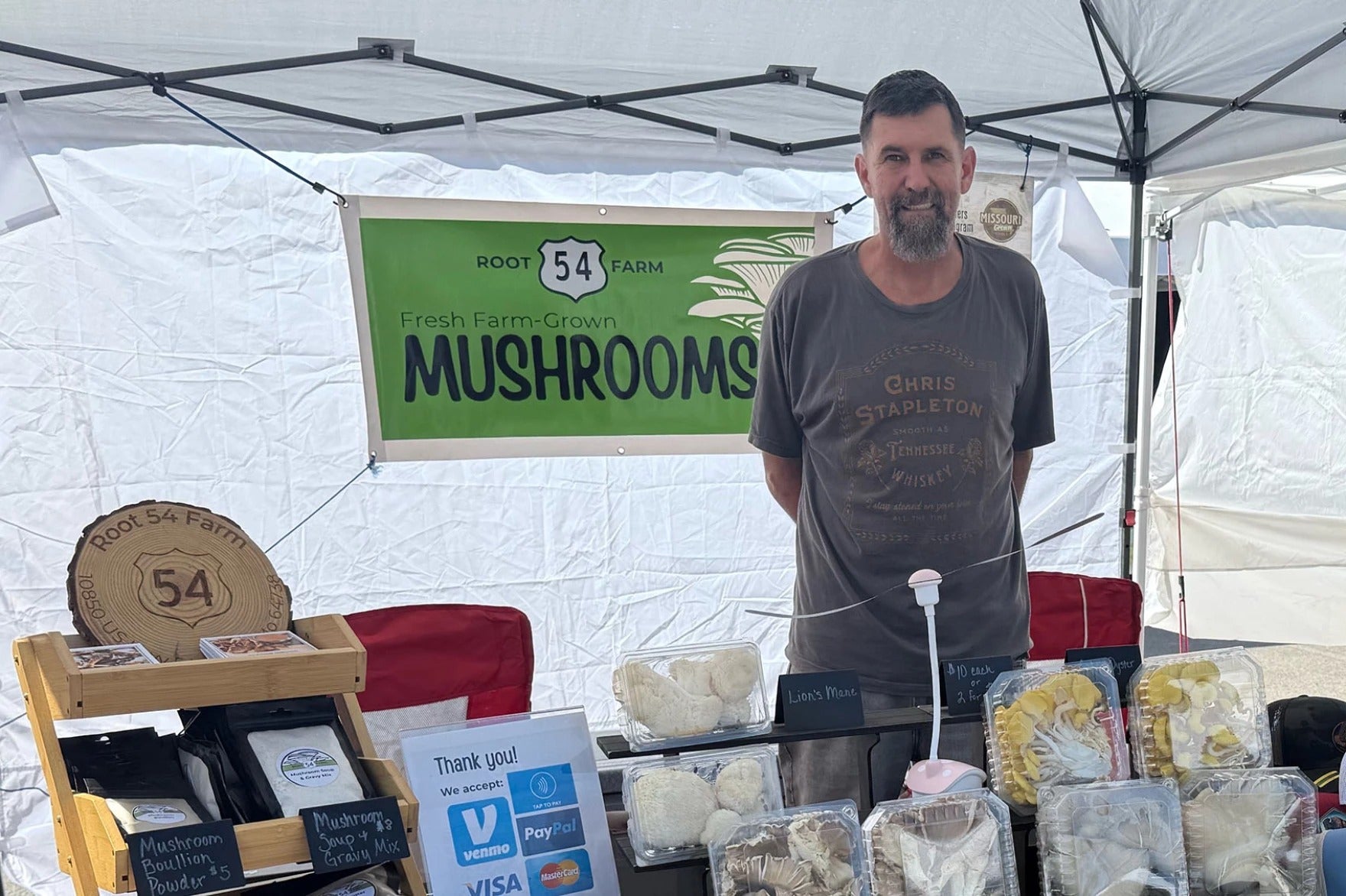A man stands behind a market stall displaying various packaged mushrooms under a Root 54 Farm Fresh Farm-Grown Mushrooms sign. Payment options are visible on the table.