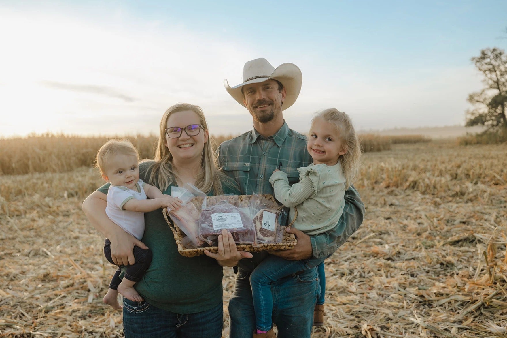 A man in a cowboy hat and a woman stand in a cornfield holding two young children and a basket of packaged food.