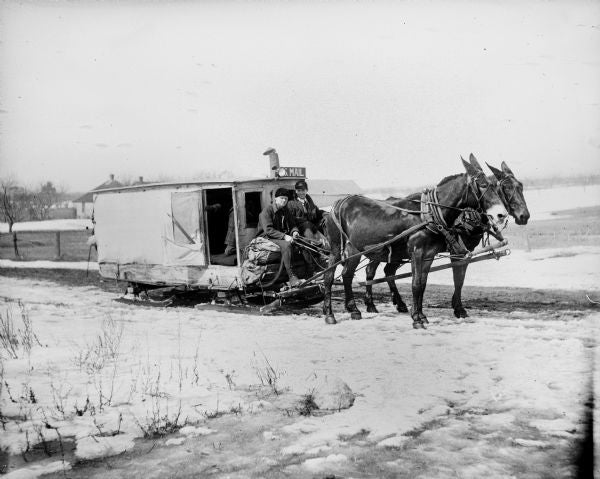 Two mules pull a sled-style mail delivery wagon with two people inside, traveling on a snowy rural road.