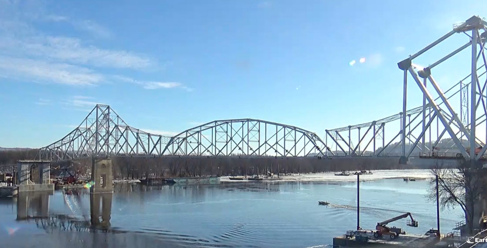 A large steel truss bridge spans a wide river, with part of the structure under construction or demolition; cranes and equipment are visible near the water.
