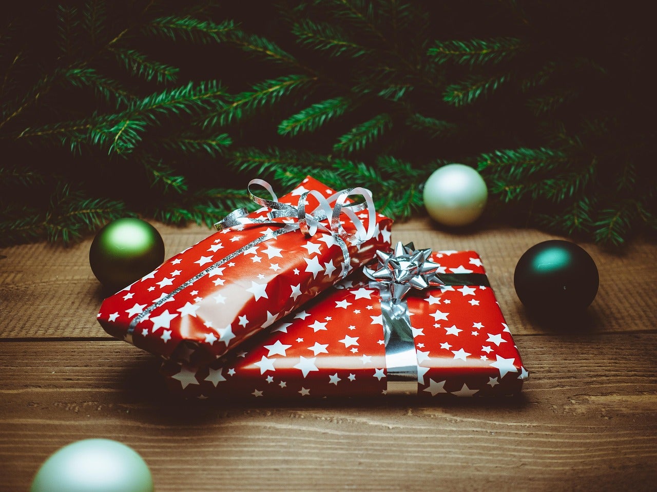 Two red and white star-patterned gift boxes with silver bows placed on a wooden surface, surrounded by Christmas tree branches and ornaments.