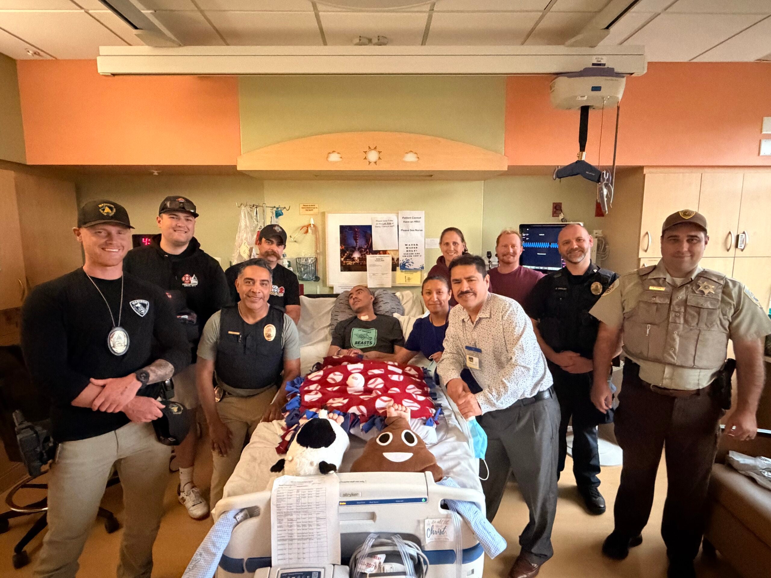 A group of law enforcement officers and hospital staff stand around a smiling patient in a hospital bed, posing together in a brightly lit patient room.
