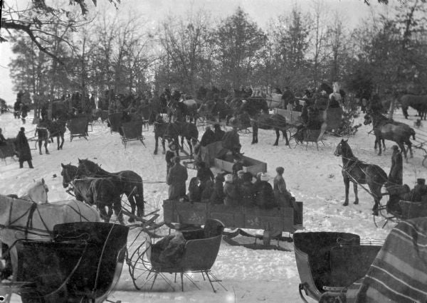 A large group of people gathered outdoors in winter, sitting in horse-drawn sleighs on a snow-covered ground with trees in the background.