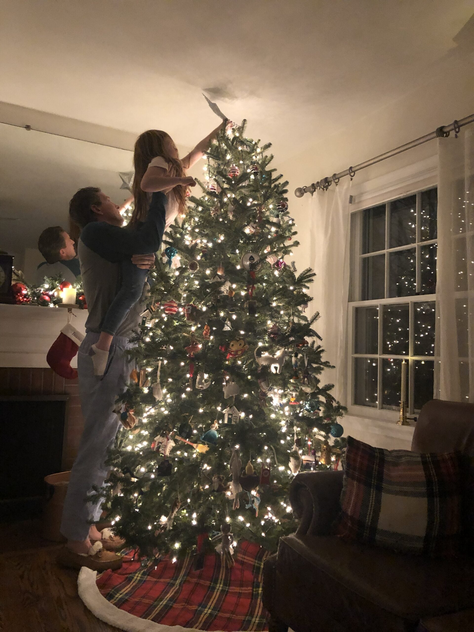 An adult lifts a child to place an ornament or decoration on the top of a lit Christmas tree in a warmly decorated living room.