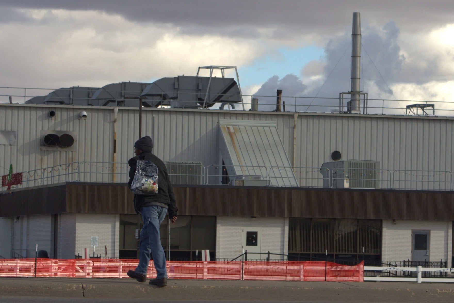 A person wearing a dark jacket and backpack walks along a sidewalk in front of an industrial building with metal vents and cloudy skies overhead.