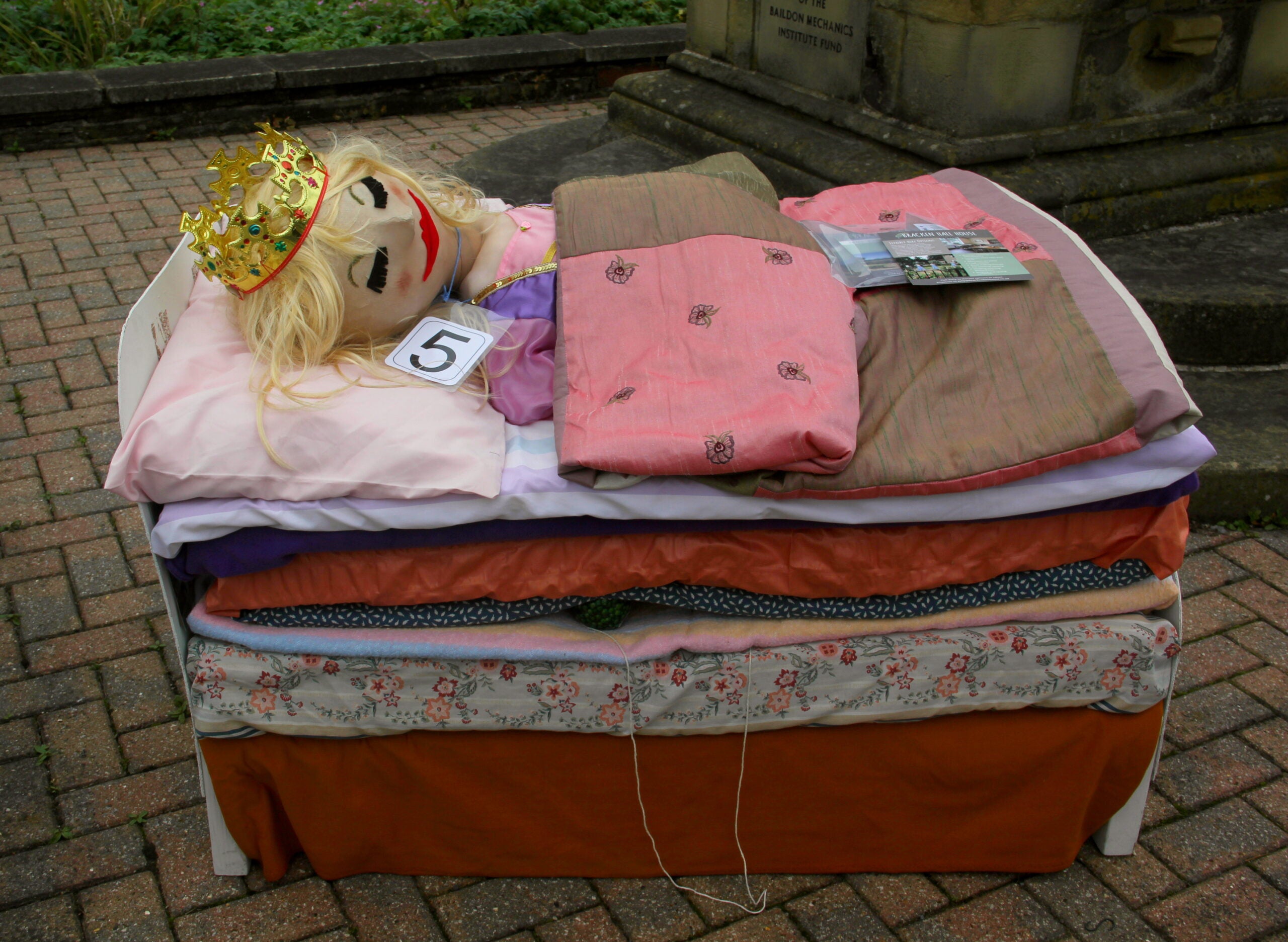 A mannequin with a crown lies under colorful blankets on a stacked mattress display outdoors, resembling the fairy tale The Princess and the Pea. A number 5 card is beside it.