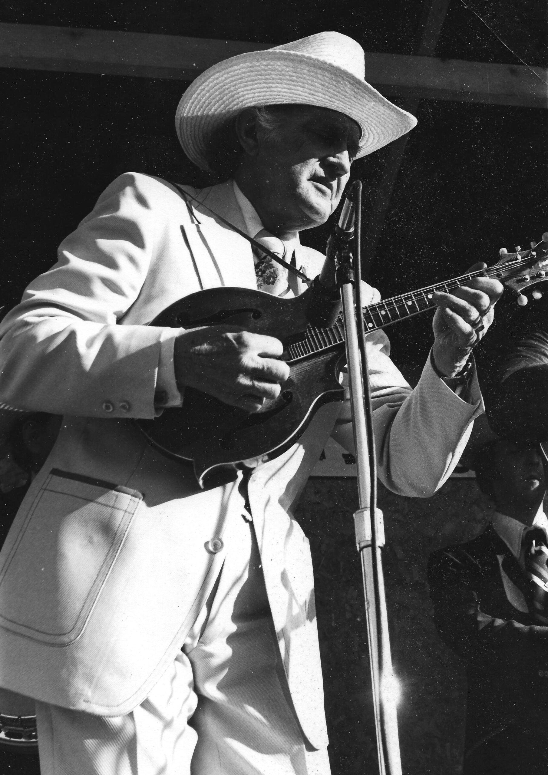 A man in a light-colored suit and wide-brimmed hat plays a mandolin on stage in front of a microphone.