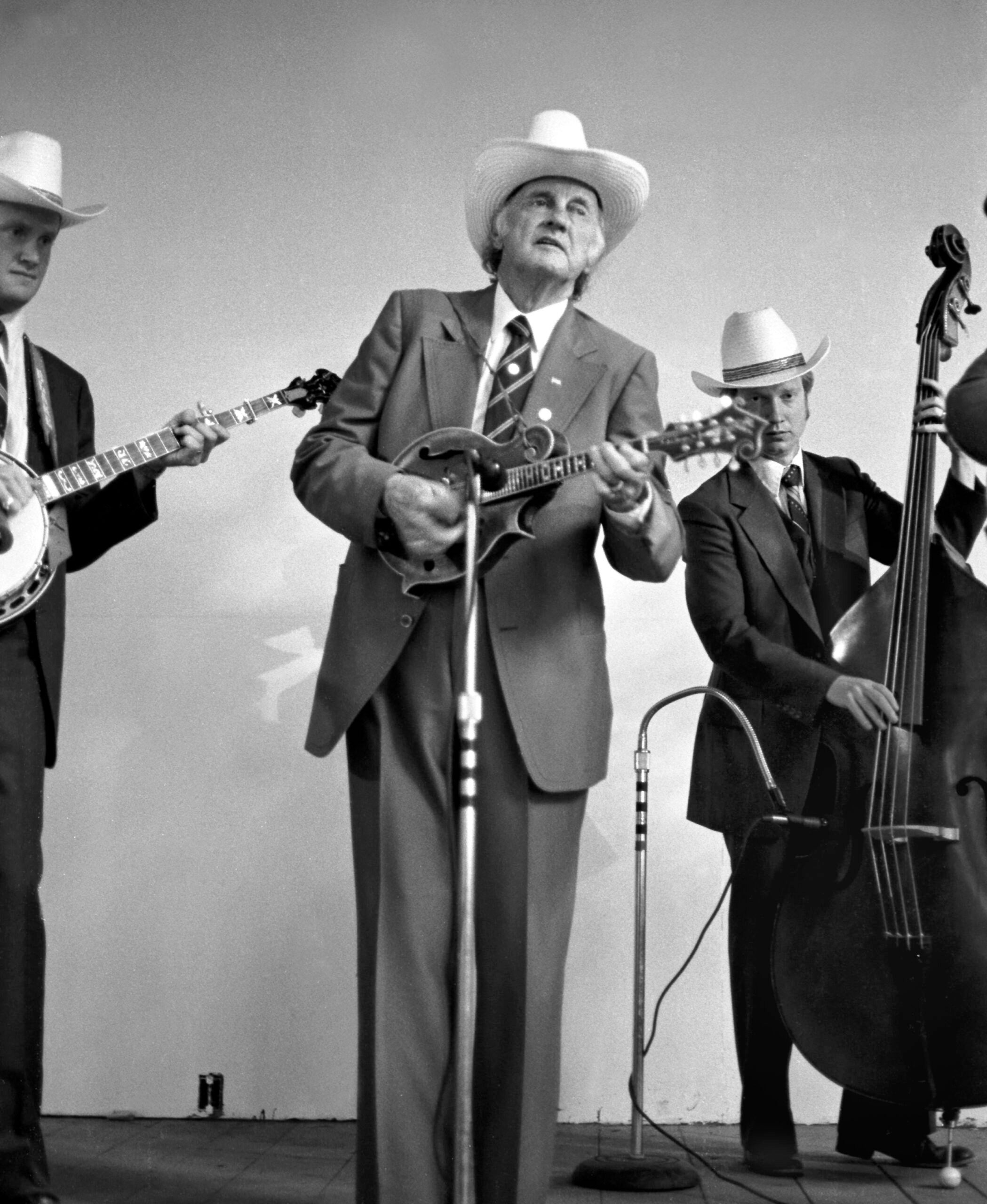 Three men in suits and cowboy hats perform on stage, playing a banjo, mandolin, and upright bass, with one singing into a microphone.