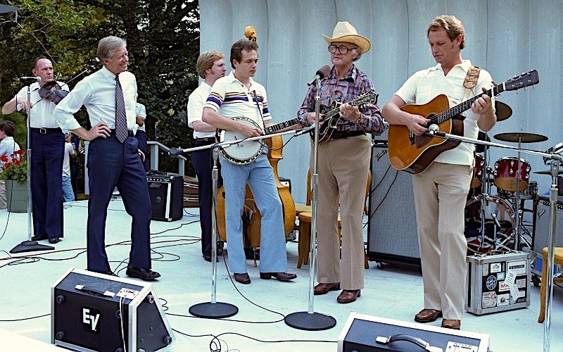 A band of six men performs on an outdoor stage with instruments including a violin, banjo, upright bass, mandolin, and guitar. They are surrounded by microphones and amplifiers.