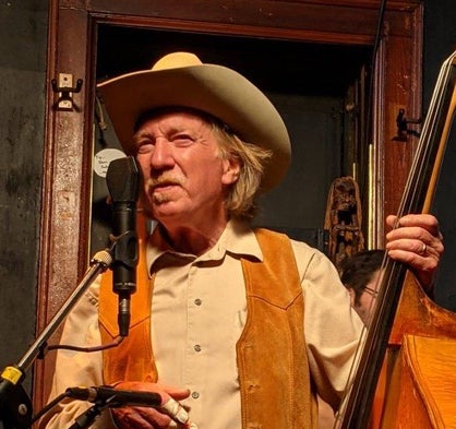 Older man in a cowboy hat and suede vest stands at a microphone, holding an upright bass, performing indoors.