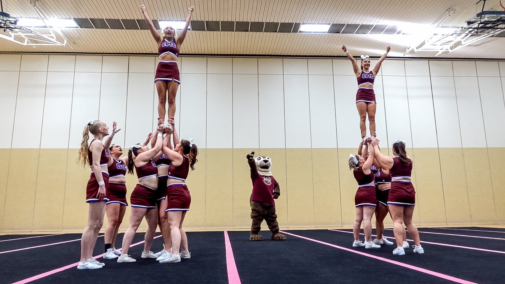 A cheerleading squad performs stunts indoors, lifting two members in the air, with a mascot standing and waving in the center.