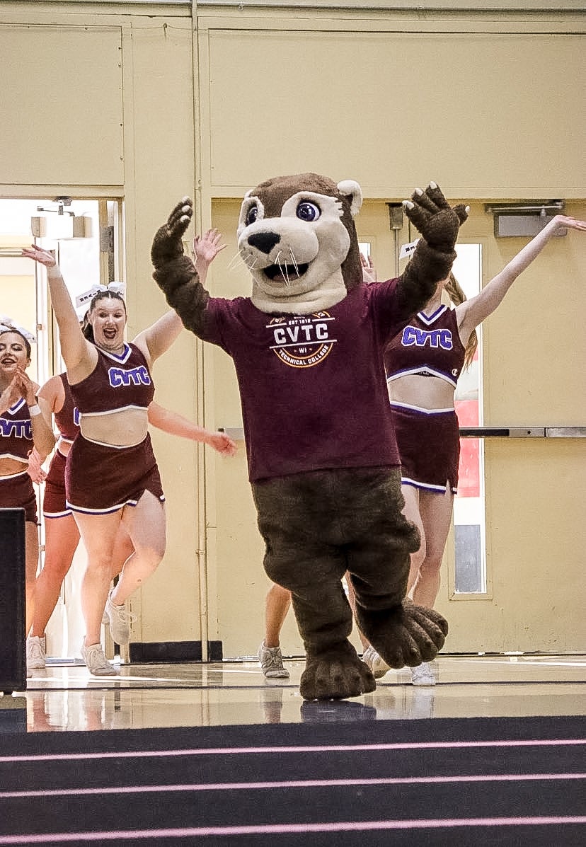 A person in an otter mascot costume leads cheerleaders onto a gym floor, all with raised arms, in front of a beige wall.